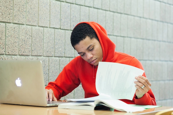 Student sitting at a table with laptop open looking at a book
